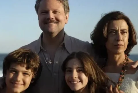 Scene from I'm Still Here, a Brazilian film featuring a family standing on the beach, with a smiling father and children, while the mother looks thoughtful.