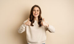 Beautiful young woman, student showing thumbs up in approval, recommending store, standing over beige background