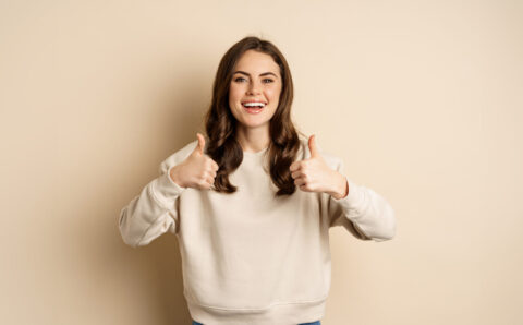 Beautiful young woman, student showing thumbs up in approval, recommending store, standing over beige background