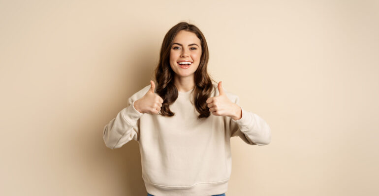 Beautiful young woman, student showing thumbs up in approval, recommending store, standing over beige background