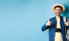 Cheerful handsome man recommending summer vacation place, showing thumbs up and smiling in approval. Tourist leave positive feedback, standing with camera and backpack on blue background