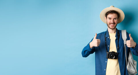 Cheerful handsome man recommending summer vacation place, showing thumbs up and smiling in approval. Tourist leave positive feedback, standing with camera and backpack on blue background