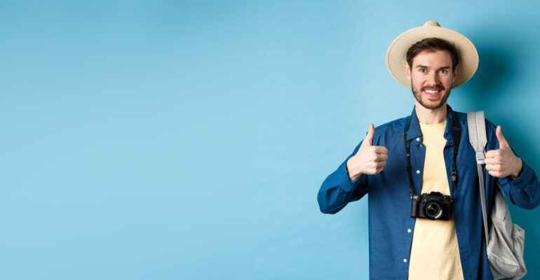 Cheerful handsome man recommending summer vacation place, showing thumbs up and smiling in approval. Tourist leave positive feedback, standing with camera and backpack on blue background