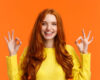 Fashion, shopping, winter holidays concept. Attractive cheerful redhead woman showing okay gestures and smiling, everything alright, approve or like product, recommend online store, orange background