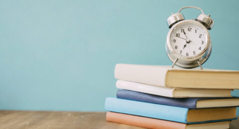 A stack of colorful books with a classic alarm clock on top, symbolizing time-efficient language learning.