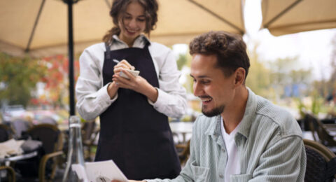 A smiling man at an outdoor restaurant ordering food from a cheerful waitress, representing a real-life dining scenario in Brazil.