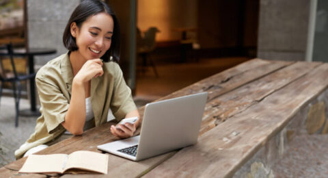 A smiling woman studying Brazilian Portuguese online, using a laptop and smartphone at an outdoor café.