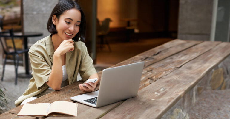 A smiling woman studying Brazilian Portuguese online, using a laptop and smartphone at an outdoor café.