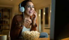 A smiling woman with headphones watching a movie on her computer while enjoying popcorn, representing a fun way to learn Portuguese.