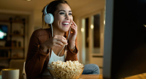 A smiling woman with headphones watching a movie on her computer while enjoying popcorn, representing a fun way to learn Portuguese.