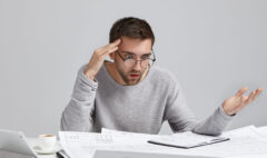 Portrait of confused male designer or architect, feels stressed, being nervous, keeps hand on head, stares in blueprint. Exhausted bearded man creates construction project alone, has some troublems