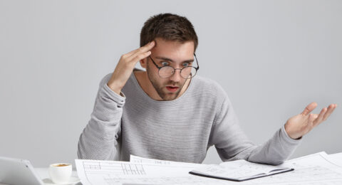 Portrait of confused male designer or architect, feels stressed, being nervous, keeps hand on head, stares in blueprint. Exhausted bearded man creates construction project alone, has some troublems