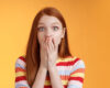 Shocked speechless impressed sensitive redhead european girl reacting stunning rumor gossiping find out secret gasping cover mouth palm stare camera astonished surprised, orange background