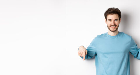 Smiling confident man pointing fingers down, showing promo banner or logo on white background, standing in casual blue sweatshirt