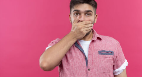 young handsome guy wearing pink polo shirt looking shocked at camera standing over pink background